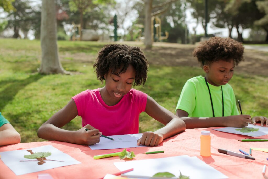 Two kids sitting down at a table doing arts and crafts. This article talks about different creative arts and craft ideas Miami summer camp organizers and counselors can use in their camps.